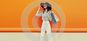 Beautiful young woman with shopping bags wearing a white striped shirt, black round straw hat on orange background