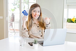 Beautiful young woman shopping online using laptop and credit card pointing and showing with thumb up to the side with happy face