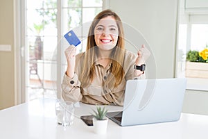 Beautiful young woman shopping online using laptop and credit card screaming proud and celebrating victory and success very