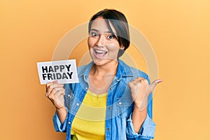 Beautiful young woman with short hair holding happy friday message paper pointing thumb up to the side smiling happy with open