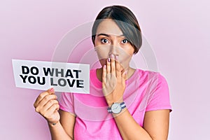 Beautiful young woman with short hair holding paper with do what you love text covering mouth with hand, shocked and afraid for