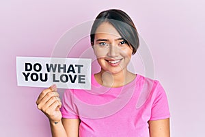 Beautiful young woman with short hair holding paper with do what you love text looking positive and happy standing and smiling