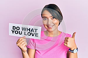 Beautiful young woman with short hair holding paper with do what you love text smiling happy and positive, thumb up doing