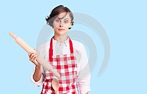 Beautiful young woman with short hair wearing professional baker apron holding kneading roll thinking attitude and sober