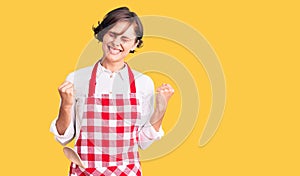 Beautiful young woman with short hair wearing professional cook apron excited for success with arms raised and eyes closed