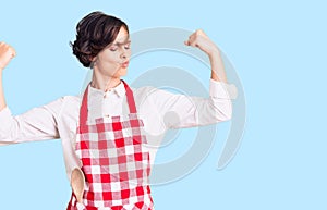 Beautiful young woman with short hair wearing professional cook apron showing arms muscles smiling proud