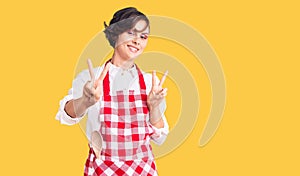 Beautiful young woman with short hair wearing professional cook apron smiling looking to the camera showing fingers doing victory