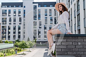 Beautiful young woman in shorts and a hat posing while sitting on a stone bench. A full-length portrait of a Caucasian