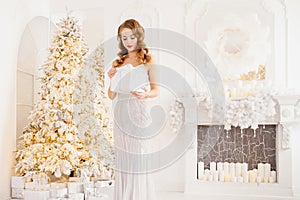 Beautiful young woman in silver dress opens gift against background of New Year tree and fireplace, white color. Dream