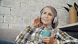 Beautiful young woman is singing listening to favourite song through headphones and holding cup of hot tea. Modern