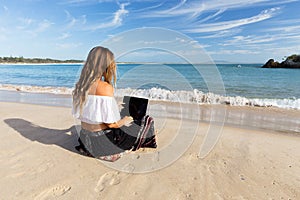 A beautiful young woman sits on the beach beside the sea and crashing waves while working remotely on a laptop