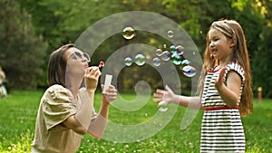 Beautiful young woman sits on green grass in the park and blows soap bubbles. Her 6-year-old daughter happily catches