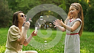 Beautiful young woman sits on green grass in the park and blows soap bubbles. Her 6-year-old daughter happily catches