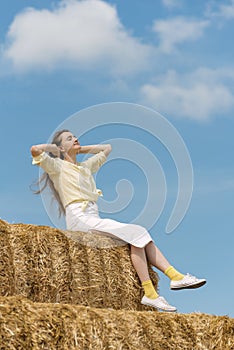 Beautiful young woman sits on haystack. Relaxing on summer field with hay stacks. Summer vacation