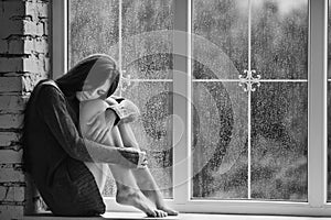 Beautiful young woman sitting alone close to window with rain drops. and sad girl. Concept of loneliness. Black