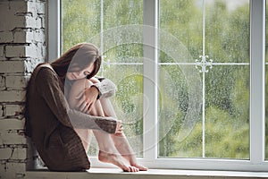 Beautiful young woman sitting alone close to window with rain drops. and sad girl. Concept of loneliness photo