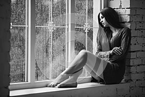 Beautiful young woman sitting alone near window with rain drops. and sad girl with long slim legs. Concept of