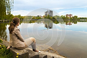 Beautiful young woman sitting on the bank of big lake with modern city skyline of buildings under construction
