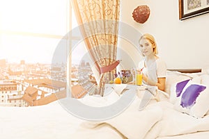 Beautiful young woman sitting on bed and having breakfast