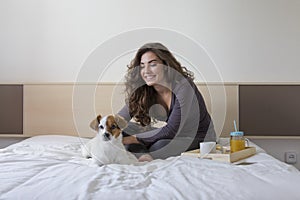 Beautiful young woman sitting on bed with her cute small dog besides. Home, indoors and lifestyle. She is having breakfast on bed