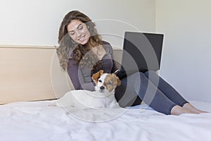 beautiful young woman sitting on bed with her cute small dog besides. She is working on laptop and smiling. Breakfast at home,