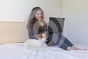 beautiful young woman sitting on bed with her cute small dog besides. She is working on laptop and smiling. Breakfast at home,