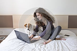 Beautiful young woman sitting on bed with her cute small dog besides. She is working on laptop and smiling. Home, indoors and