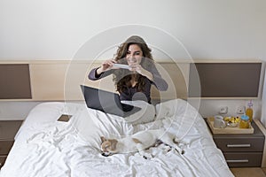 Beautiful young woman sitting on bed with her cute small dog besides. She is working on laptop and taking a picture of her dog