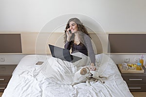 beautiful young woman sitting on bed with her cute small dog besides. She is working on laptop and talking on her mobile phone.