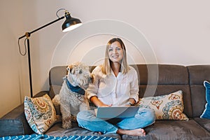 Beautiful young woman sitting on couch in living room with laptop surfing Internet. Woman with pet dog chatting with friend in