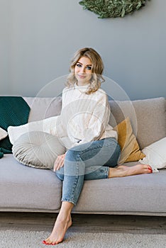 Beautiful young woman is sitting on a cozy sofa with pillows in a room decorated for the new year or Christmas