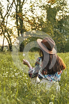 Beautiful Young Woman sitting on the field in green grass and blowing dandelion. Outdoors. Enjoy Nature. Healthy Smiling Girl on