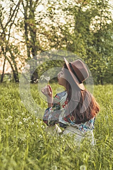 Beautiful Young Woman sitting on the field in green grass and blowing dandelion. Outdoors. Enjoy Nature. Healthy Smiling Girl on