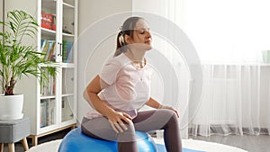 Beautiful young woman sitting on fitball and doing fitness exercises at home. Concept of healthcare, sports and yoga at home