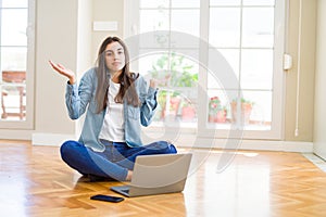 Beautiful young woman sitting on the floor with crossed legs using laptop clueless and confused expression with arms and hands