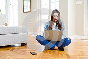 Beautiful young woman sitting on the floor with crossed legs using laptop scared in shock with a surprise face, afraid and excited