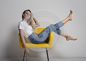 Beautiful young woman is sitting or lying on yellow chair with legs up and looking arrogantly at camera