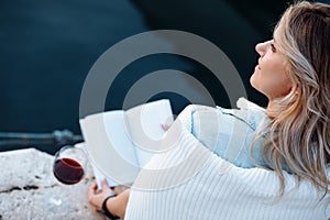 beautiful young woman is sitting on pier with glass of red wine and book having great time on vacation , happy evening.