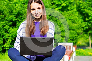 Beautiful young woman sitting in public park and working on laptop computer. Portrait of smiling woman studying on laptop at wile