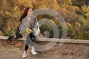 Beautiful young woman sitting on the roadside on nature background and thinking about life. Bouquet of autumn leaves in her hands