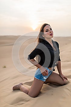Beautiful young woman sitting sand while sitting on desert dune during sunset