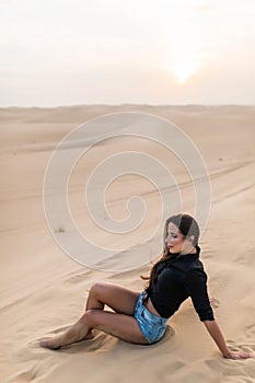 Beautiful young woman sitting sand while sitting on desert dune during sunset