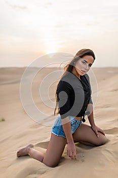 Beautiful young woman sitting sand while sitting on desert dune during sunset