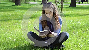 Beautiful young woman sitting in a summer park on the grass and reading a book of the Bible
