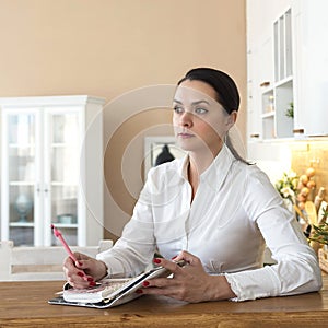 Beautiful young woman sitting at the table in the kitchen and taking notes