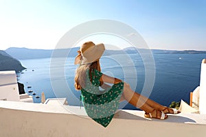 Beautiful young woman sitting on wall looking at stunning view of Mediterranean sea and Caldera in Santorini, Greece, Europe.