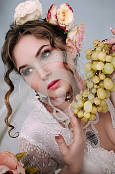 Beautiful young woman sitting on white bed and eating grapes, wearing white lace dress, room decorated with flowers. Perfect