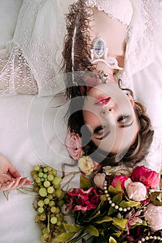 Beautiful young woman sitting on white bed and eating grapes, wearing white lace dress, room decorated with flowers. Perfect