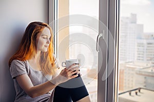 Beautiful young woman sitting on windowsill overlooking the metropolis, drinking tea or coffee from cup. Female portrait on the