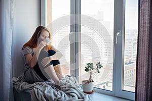 Beautiful young woman sitting on windowsill overlooking the metropolis, drinking tea or coffee from cup. Female portrait on the